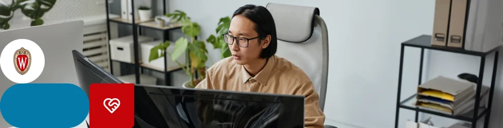 A person with obscured face working at a desk with multiple computer screens and a laptop with stickers.